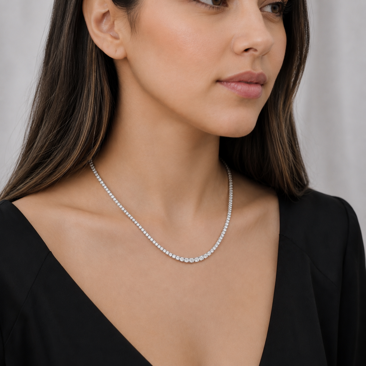 A close-up of a woman wearing a delicate silver tennis necklace with small round-cut stones, styled with a black V-neck top against a soft light gray background.