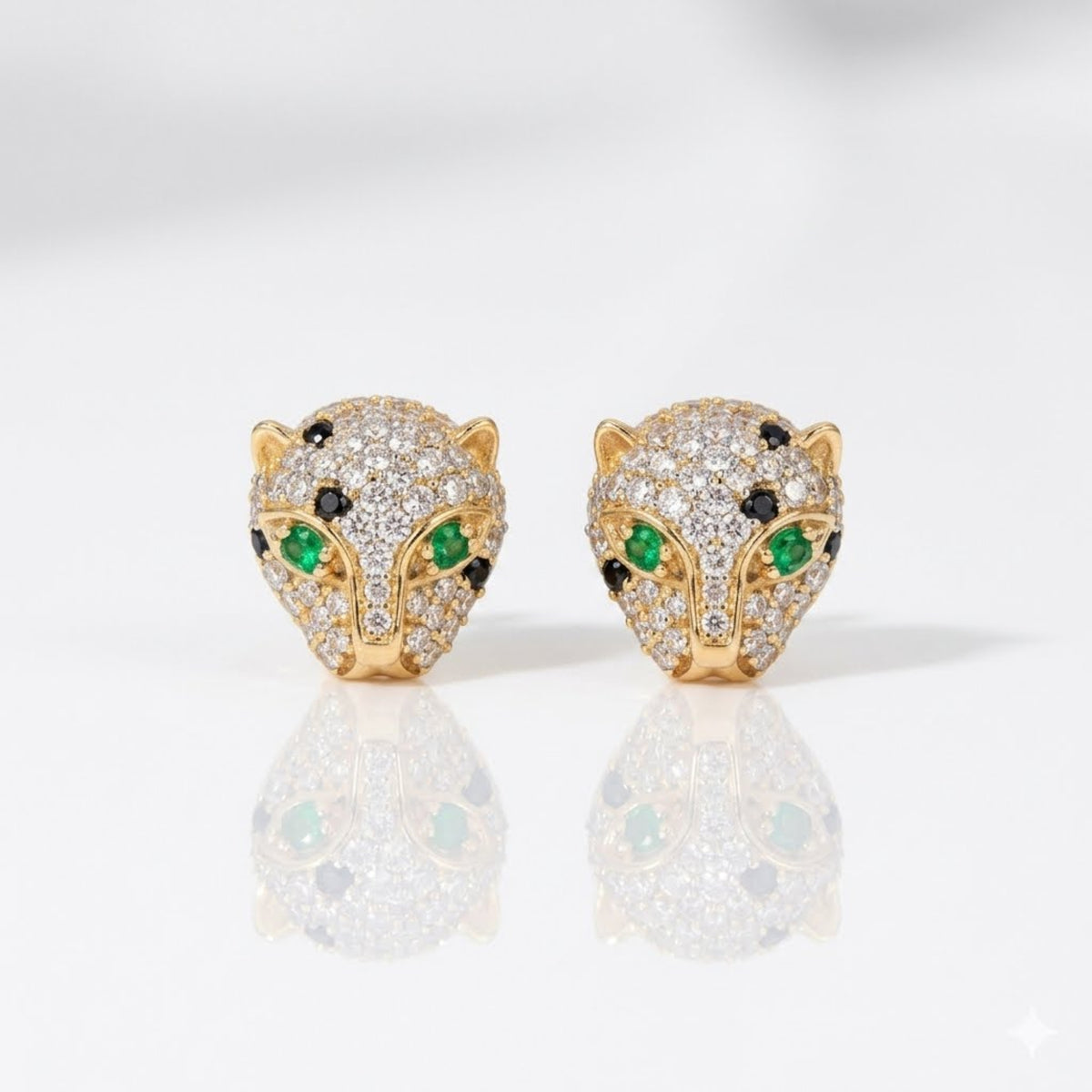 A pair of gold panther head stud earrings paved with clear diamonds, featuring green and black gemstone eyes, displayed on a reflective white surface.