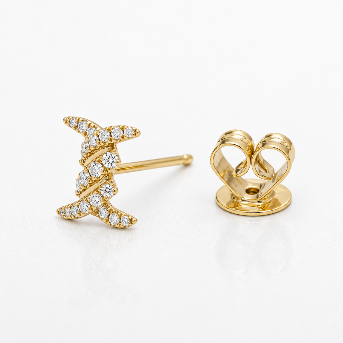 A pair of gold stud earrings on a clean white background. One earring is shown from the front, featuring a twisted, diamond-encrusted crescent or leaf-like design, while the other is shown from the back, displaying a polished gold heart-shaped post and secure backing.