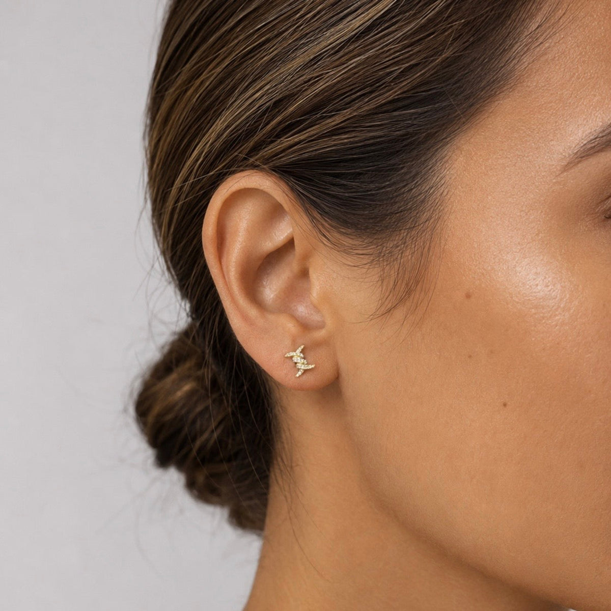 A close-up of a woman's ear wearing a small gold earring with a stylized abstract symbol, set against a neutral light gray background.