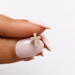 A close-up of a hand with pale pink manicured nails holding a small gold cross stud earring set with diamonds against a clean white background.