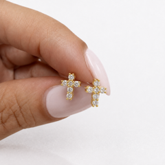 A close-up of a hand with pale pink manicured nails holding two small gold cross stud earrings set with diamonds against a clean white background.