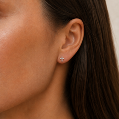 A close-up of a woman's ear wearing a small cross stud earring with tiny diamond stones.