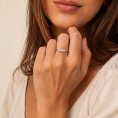 A close-up lifestyle shot of a woman wearing a delicate silver ring with small clear diamonds on her ring finger. The background is a soft, warm beige tone.