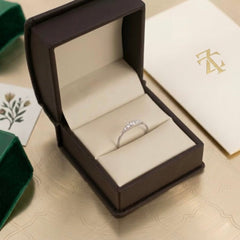 A silver ring with small clear diamonds set in a dark brown jewelry box with cream velvet lining, styled on a beige surface with branded packaging nearby.