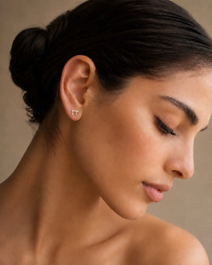 A close-up portrait of a woman with dark hair in a bun, wearing a small silver Chai stud earring. The background is a soft, neutral beige studio backdrop.