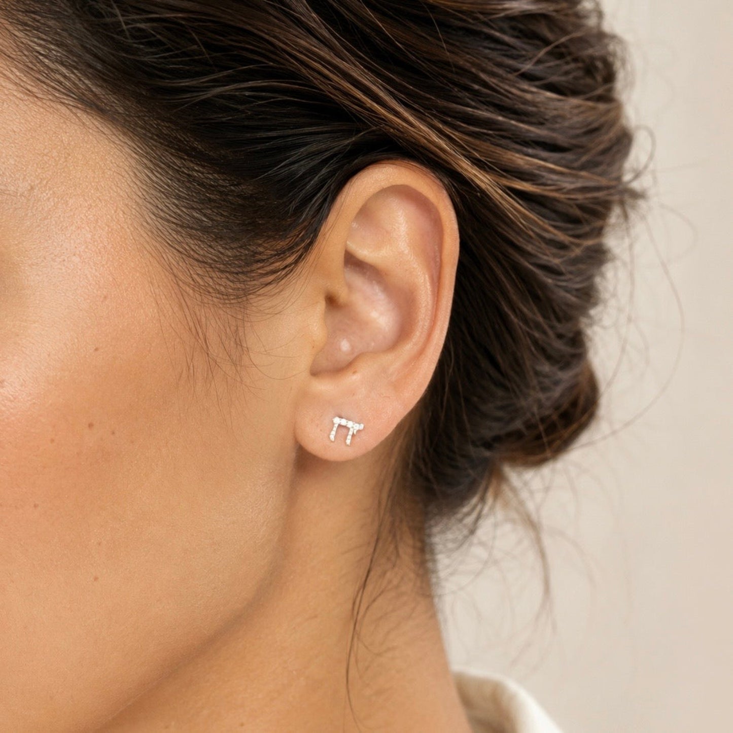A close-up of a woman's ear wearing a small silver stud Chai earring, against a soft beige background.