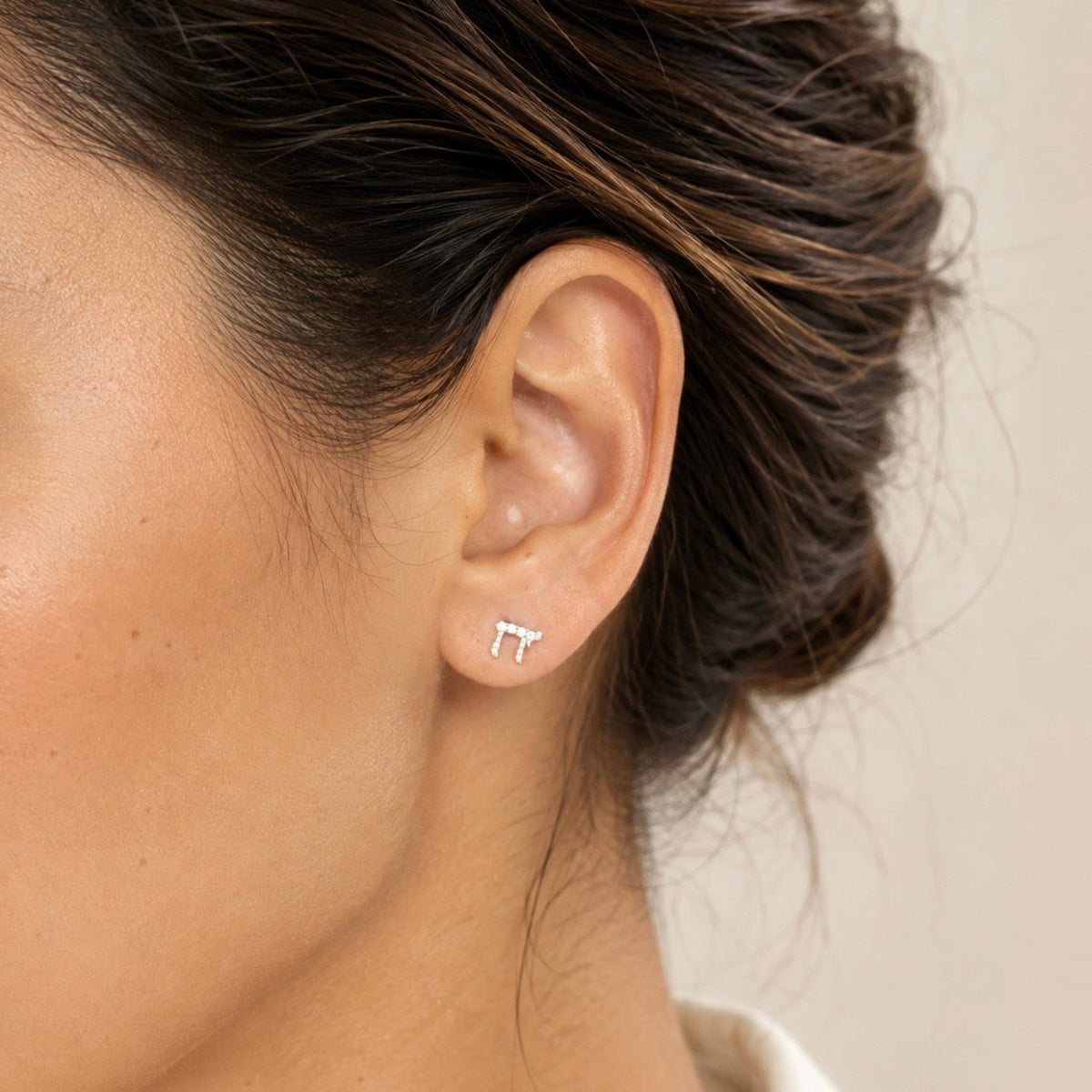A close-up of a woman's ear wearing a small silver stud Chai earring, against a soft beige background.