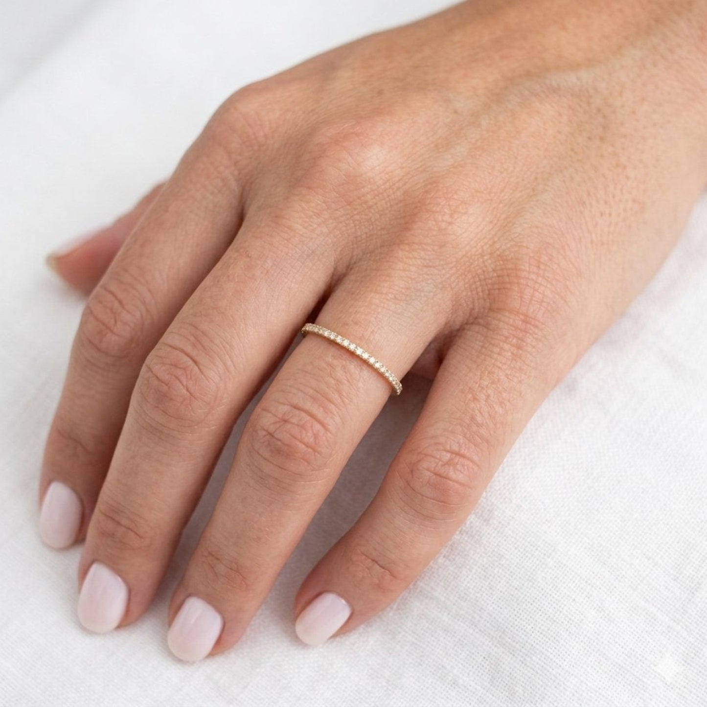 A delicate gold ring with a row of small diamonds worn on a person's ring finger, shown against a soft white textured fabric background.