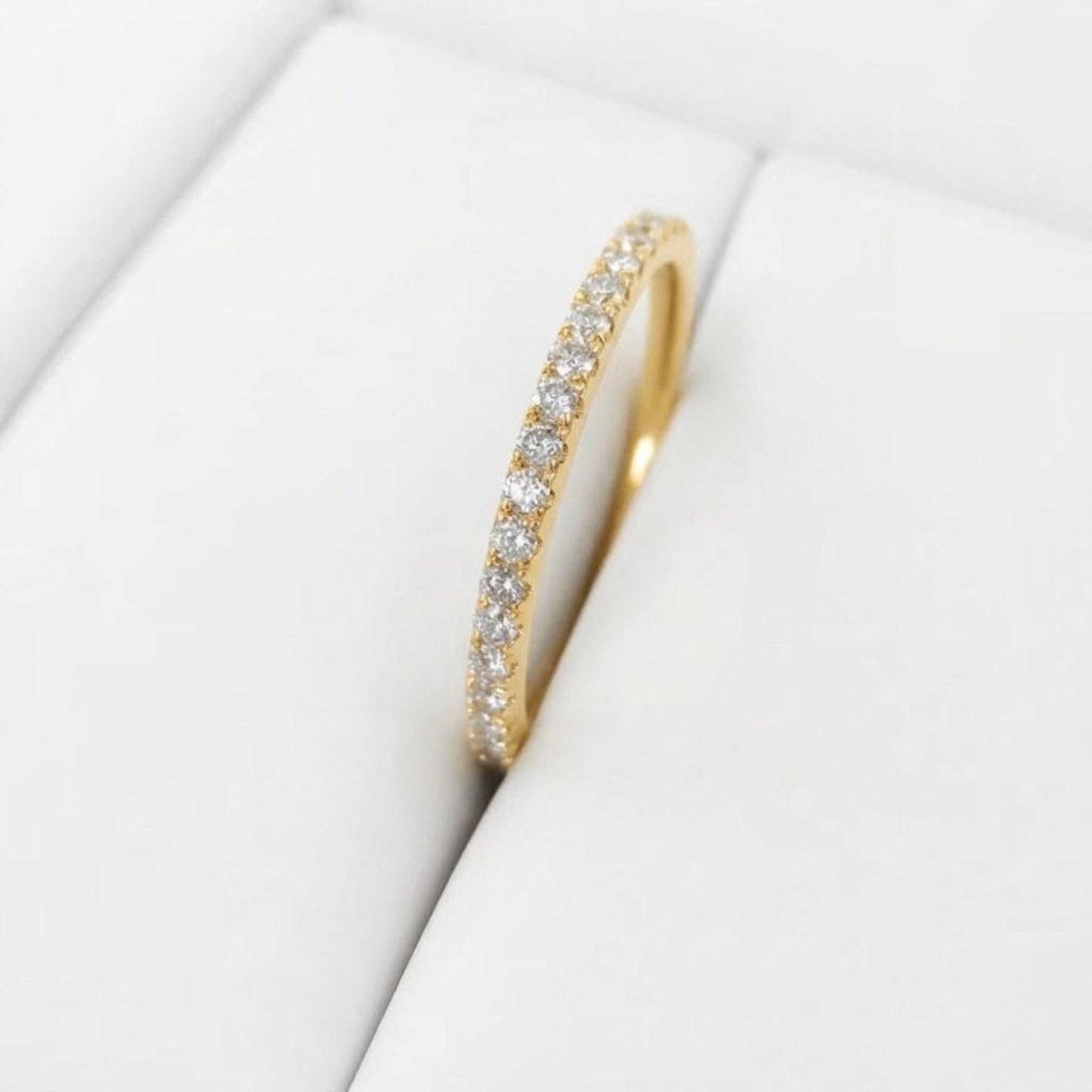 A yellow gold eternity ring set with small round-cut clear diamonds, displayed on a white textured jewelry stand.