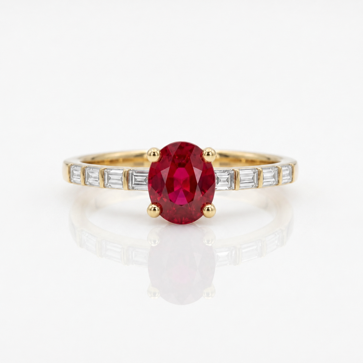 A yellow gold ring featuring an oval-cut red ruby center stone, accented by baguette-cut clear diamonds along the shoulders, set against a clean white background with a soft reflection.