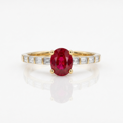 A yellow gold ring featuring an oval-cut red ruby center stone, accented by baguette-cut clear diamonds along the shoulders, set against a clean white background with a soft reflection.