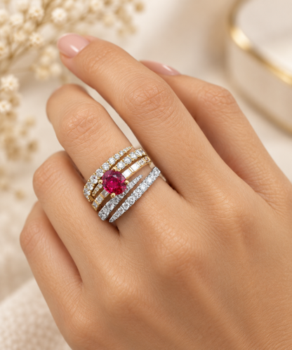 A close-up of a hand wearing a stack of three rings. The center ring features a round-cut red ruby gemstone surrounded by yellow gold and white diamond pavé bands.
