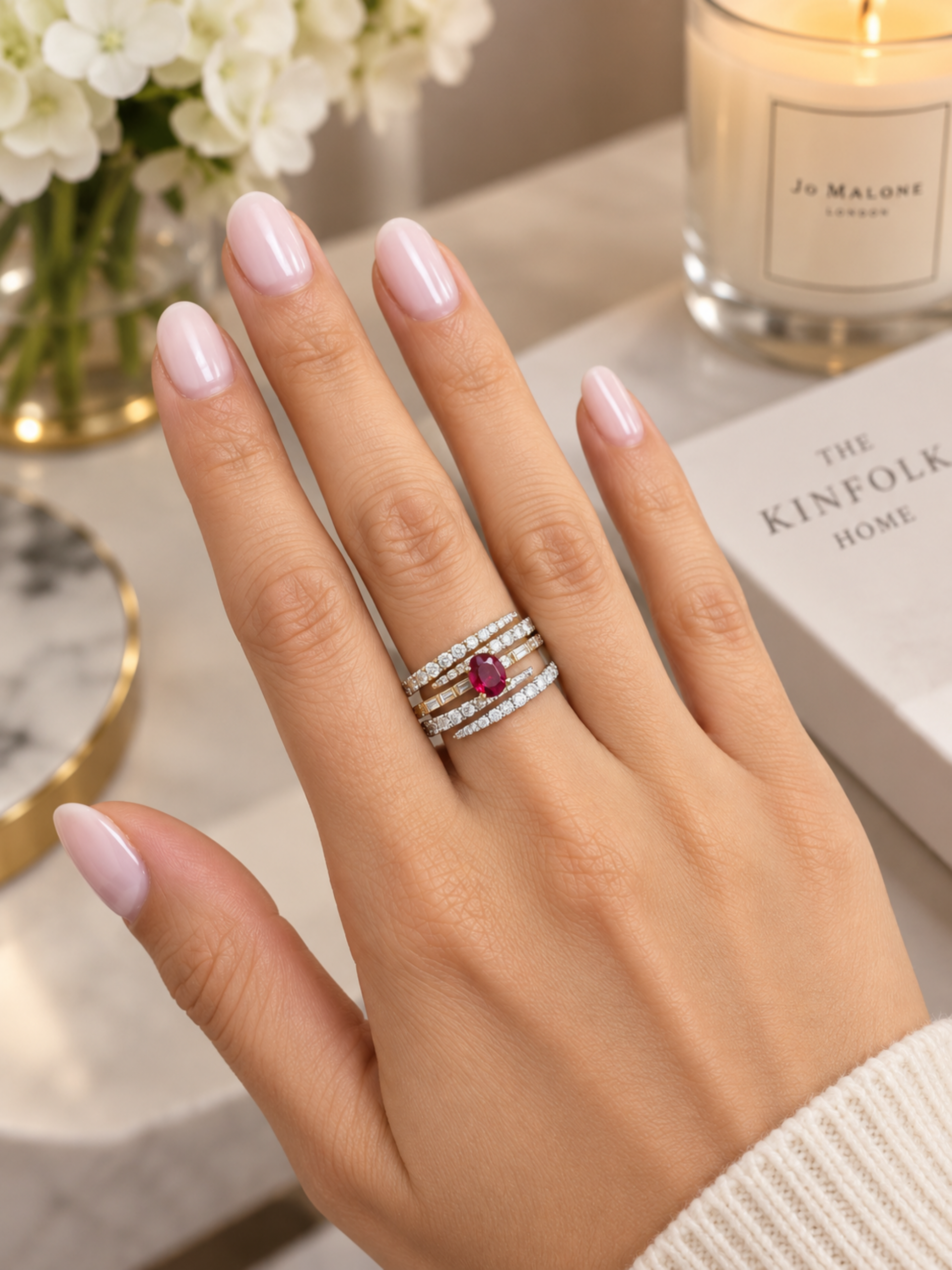 A close-up of a hand wearing a multi-band ring with a central round ruby gemstone and diamond accents, styled with a Jo Malone London candle and a book in the background.