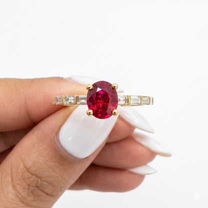 A gold ring with a large oval red ruby and baguette diamonds side stones, held between fingers with white manicured nails against a plain white background.