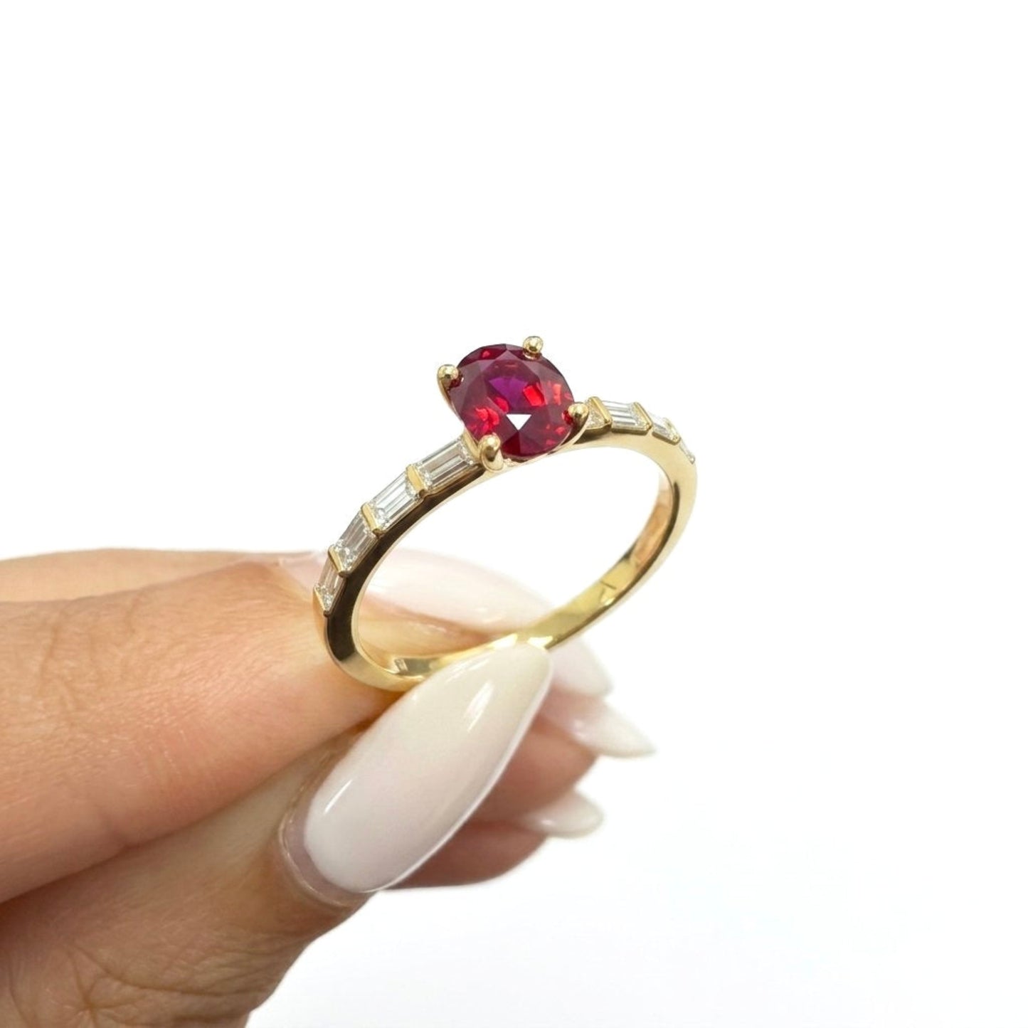 A gold ring with an oval red ruby and baguette diamonds side stones, held between fingers against a white background.