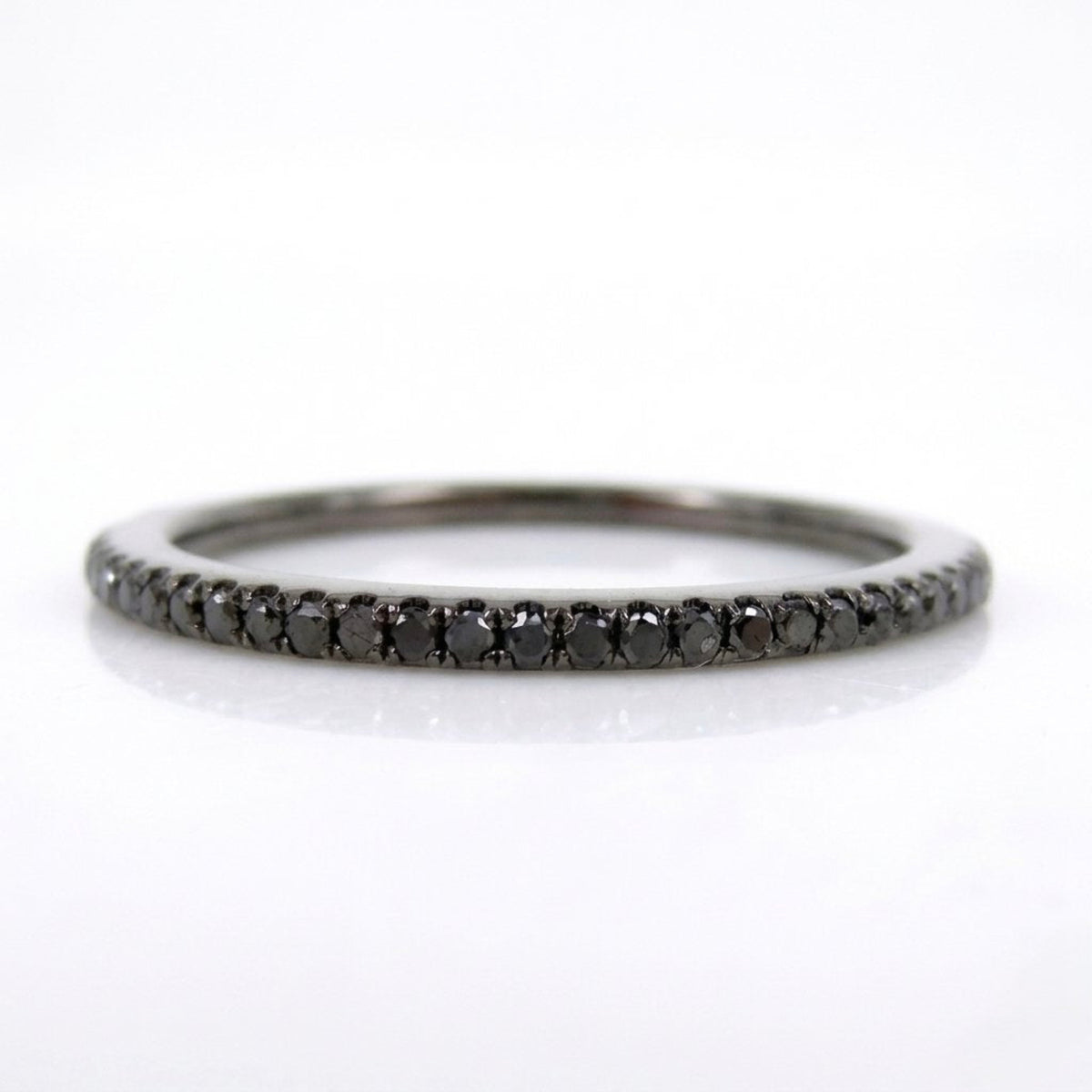 A slim black diamond eternity band ring set in a polished silver-toned metal, shown on a clean white background with a soft reflection.