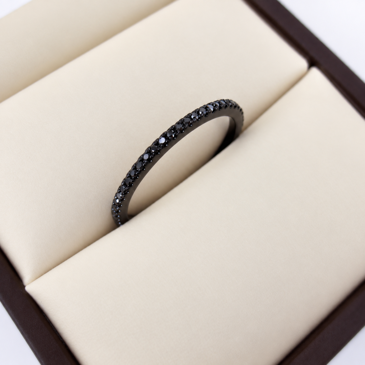 A black metal eternity ring set with small black diamonds, displayed inside a cream-colored jewelry box.