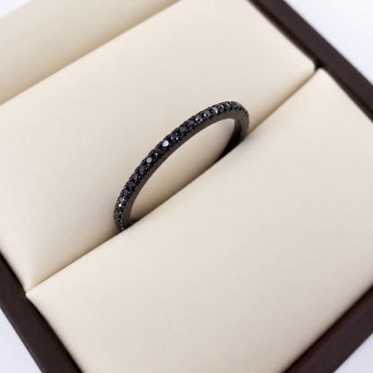 A black metal eternity ring set with small black diamonds, displayed inside a cream-colored jewelry box.