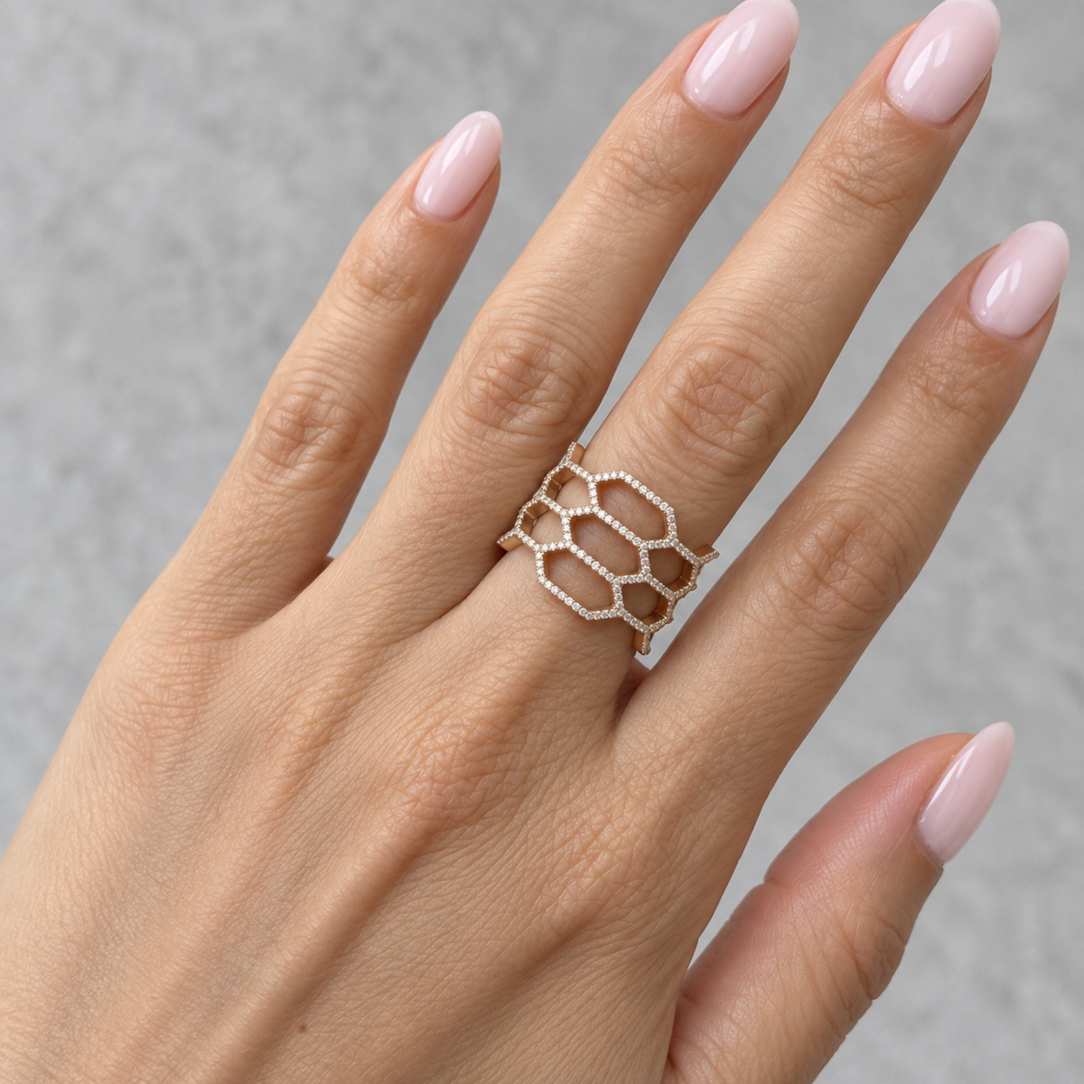 A close-up of a hand wearing a delicate honeycomb-patterned ring with small diamonds on a light gray textured background.