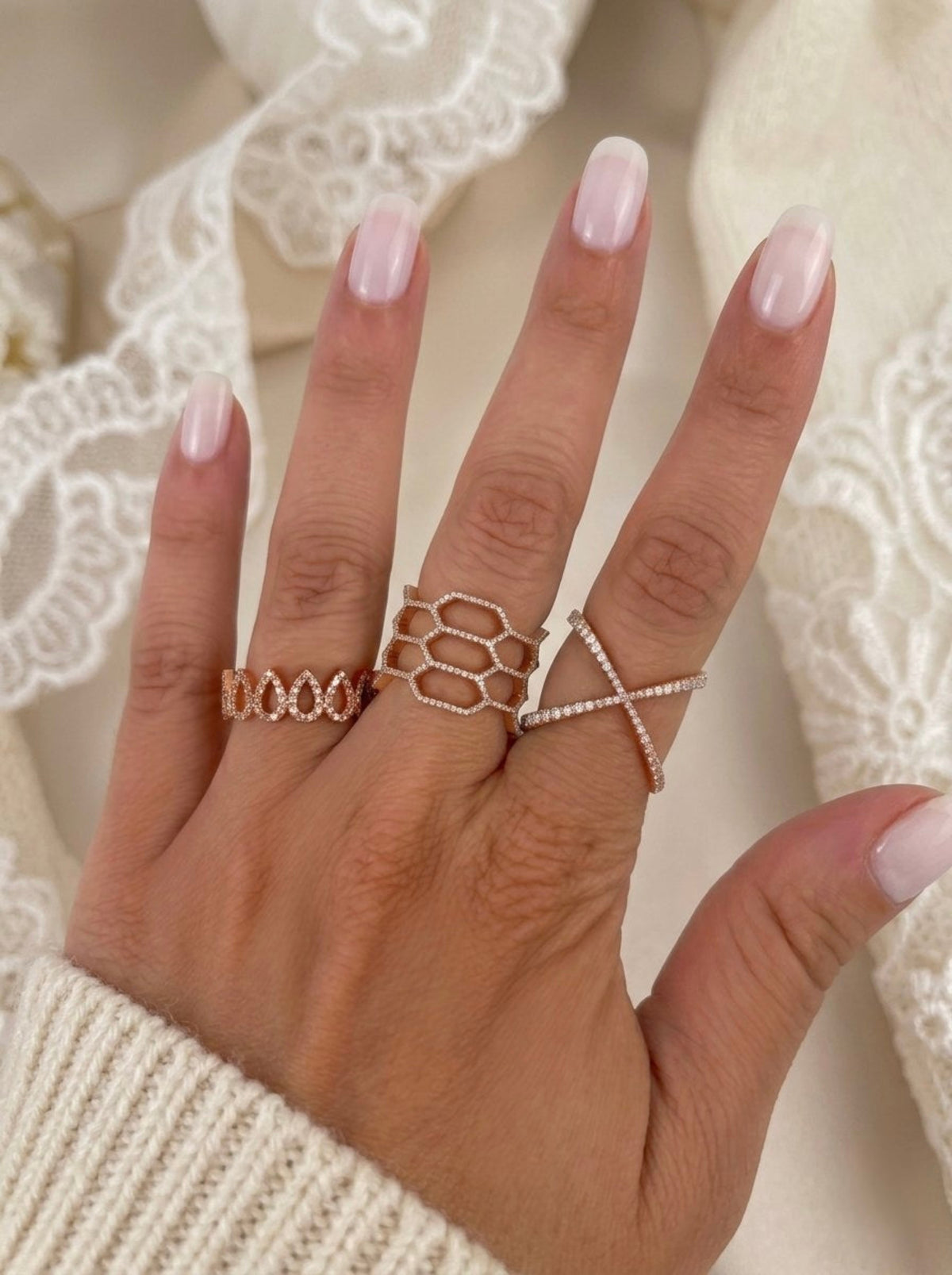A close-up of a hand wearing three rose gold rings: a textured band, a honeycomb-style ring, and an X-shaped ring with small diamonds, set against a soft cream lace background.