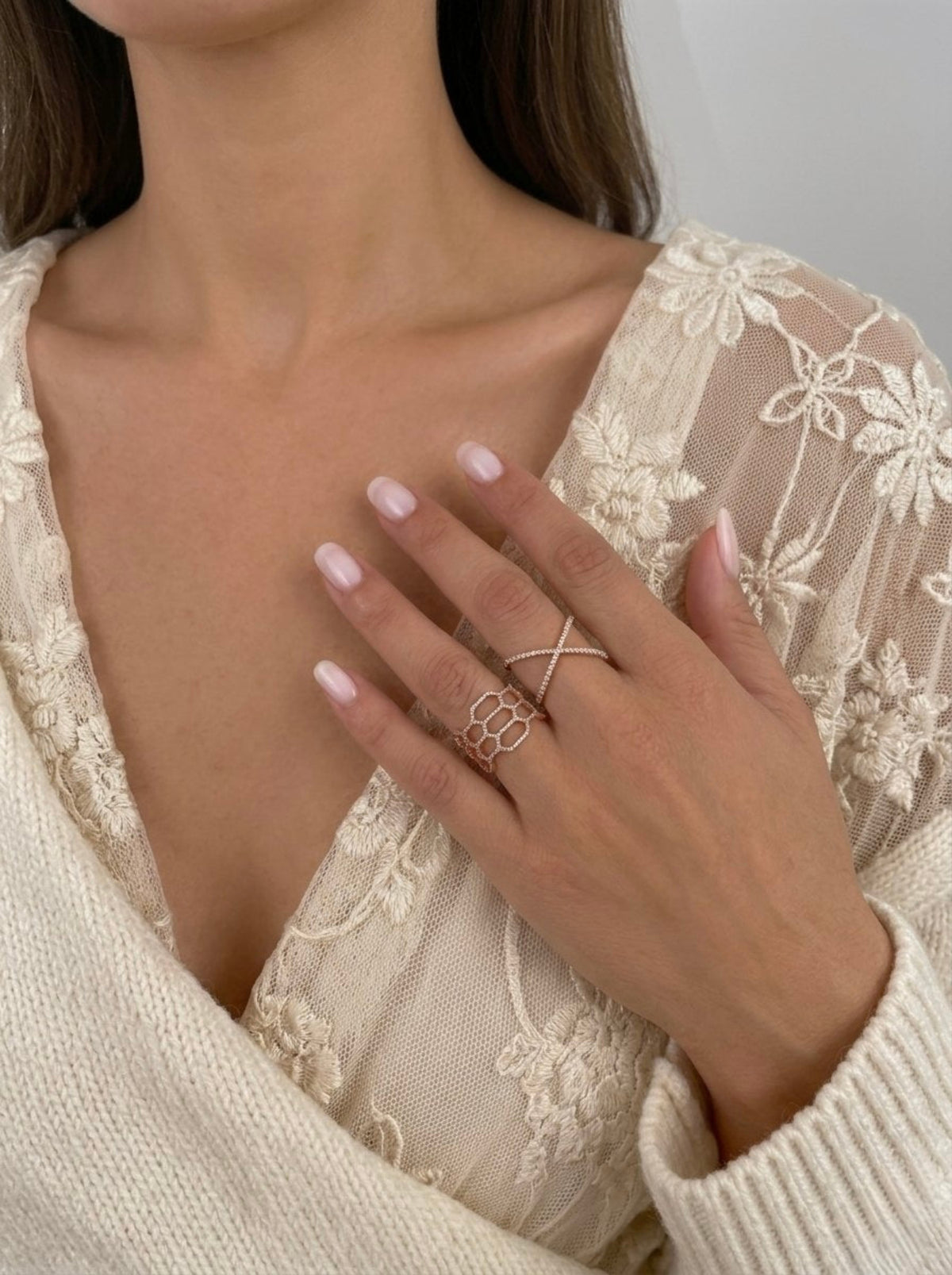 A close-up of a model's hand wearing a rose gold ring set with a small cross and a textured geometric ring, styled with a cream lace top.