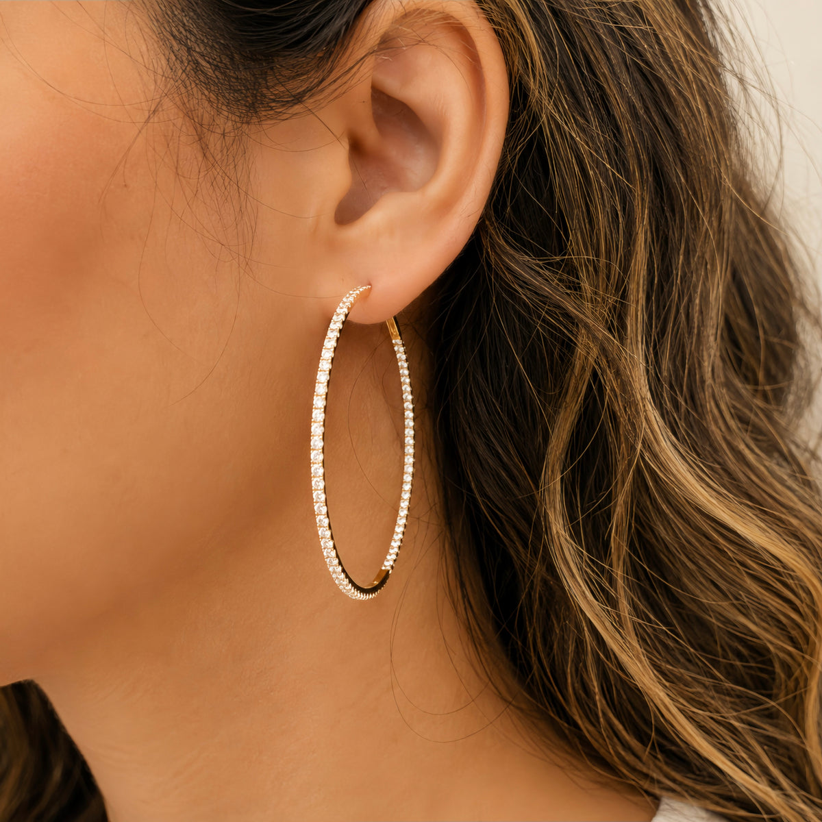 A close-up of a model's ear wearing a large, double-row hoop earring made of small clear diamonds in a gold-tone setting.