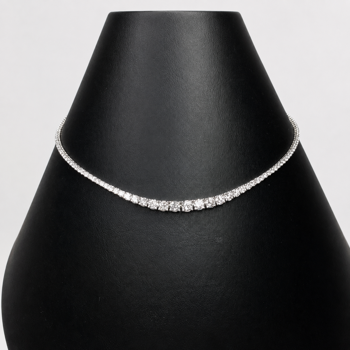A delicate silver tennis necklace with clear diamonds, displayed on a black jewelry bust against a light gray background.