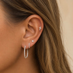 A close-up of a woman's ear wearing multiple silver-toned earrings, including a small cross stud in the lobe, a small round stud, a thin hoop, and a larger hoop set with diamonds, against a soft beige background.