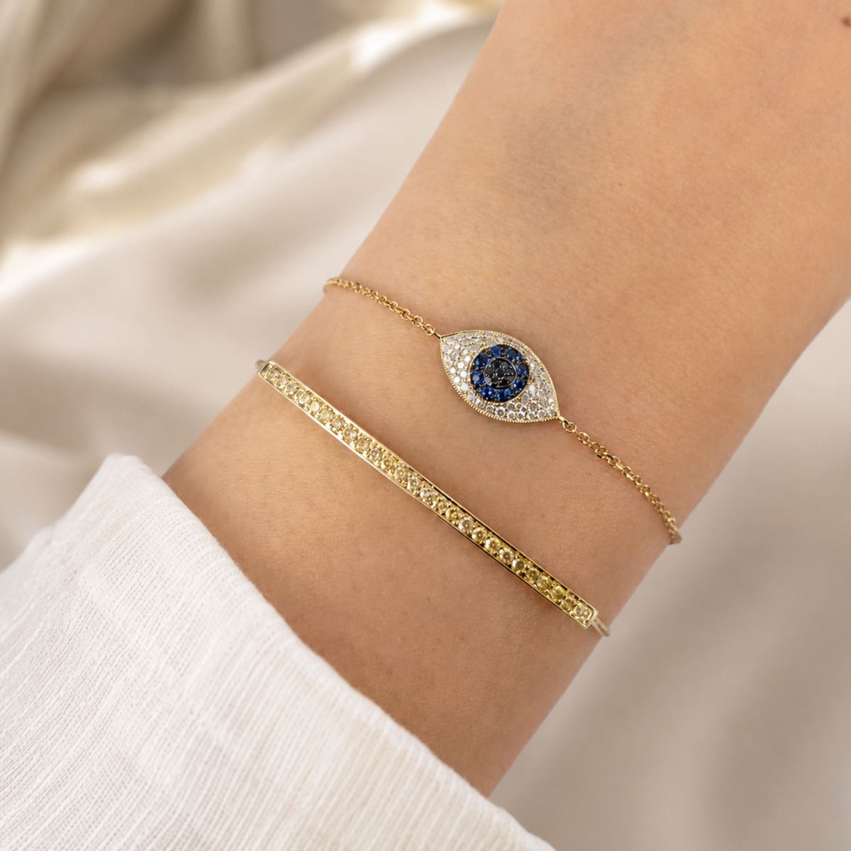 A close-up of a person's wrist wearing two delicate bracelets: a gold chain bracelet with an eye-shaped blue stone and clear diamonds halo, and a textured gold bar bracelet, against a soft cream fabric background.