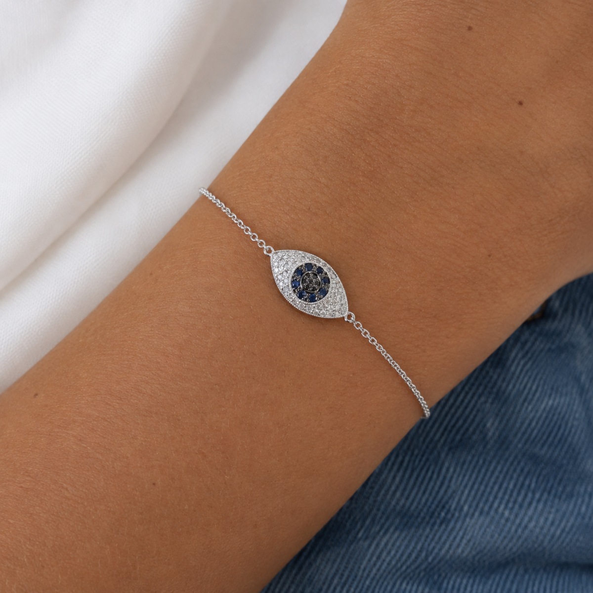 A close-up of a delicate silver chain bracelet with an evil eye charm worn on a person's wrist. The charm features a dark blue central circle surrounded by small clear diamonds, set against a background of white fabric and blue denim.