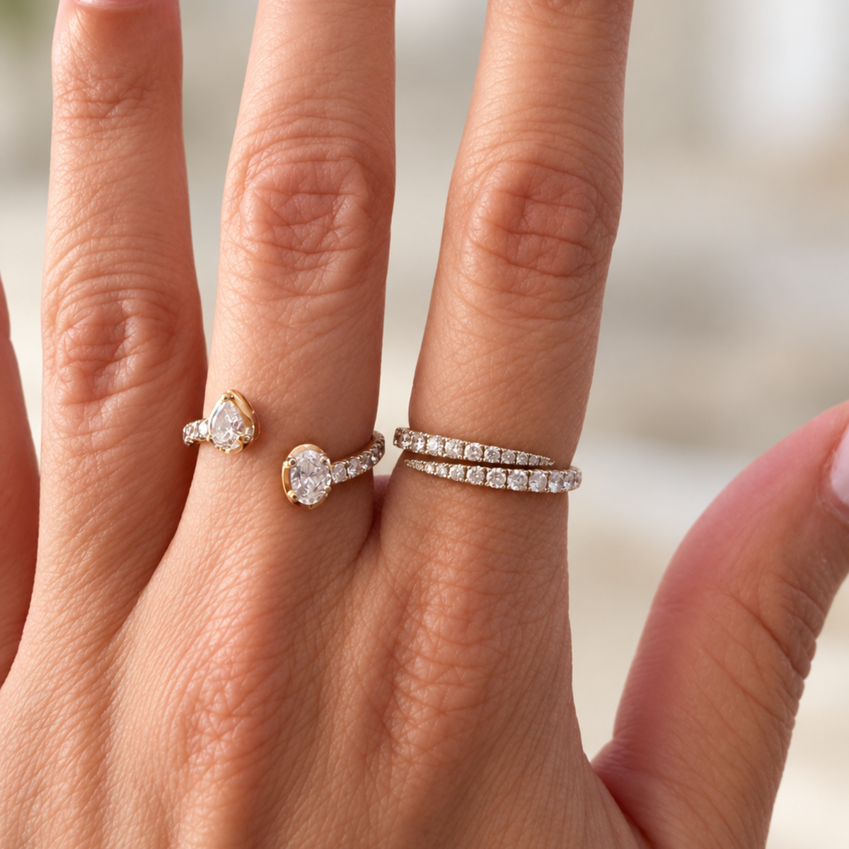 A close-up of a hand wearing three rings: a yellow gold ring with a pear-cut diamond, a white gold ring with a oval-shaped diamond, and a thin white gold diamond band wrapped around the middle finger.