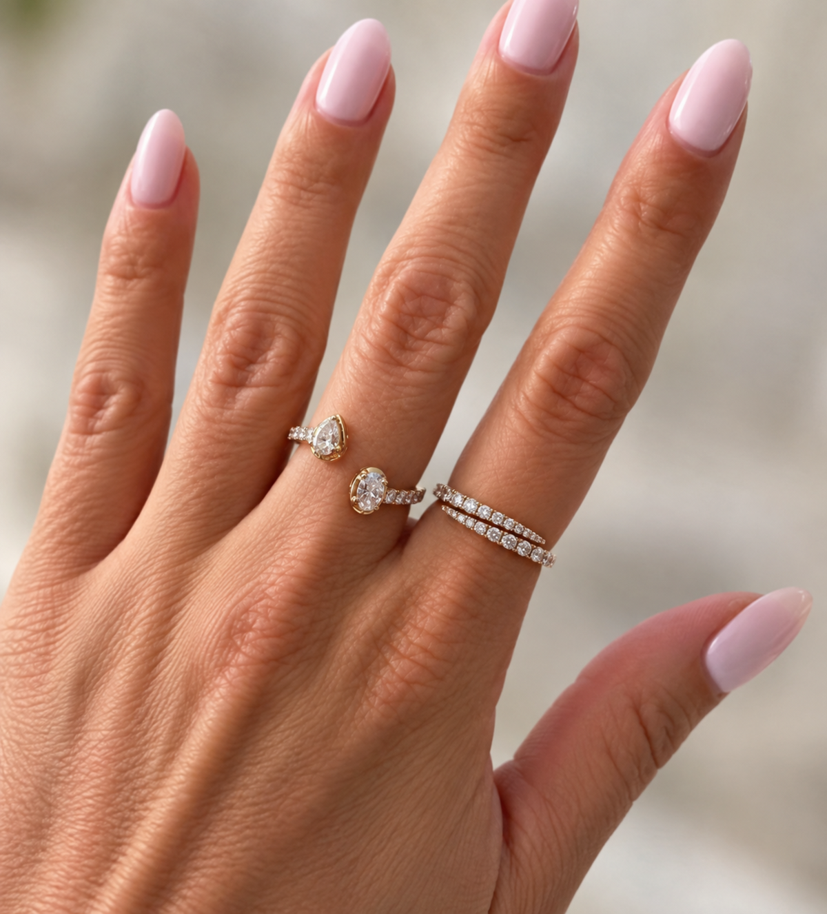 A close-up of a hand with light pink manicured nails wearing two delicate rings: a gold ring with a pear-cut clear gemstone and a silver-toned pavé band ring with small clear stones.