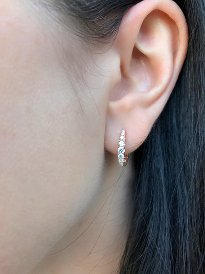 A close-up of a person's ear wearing a small vertical huggie hoop earring set with clear stones in a gold-tone setting.