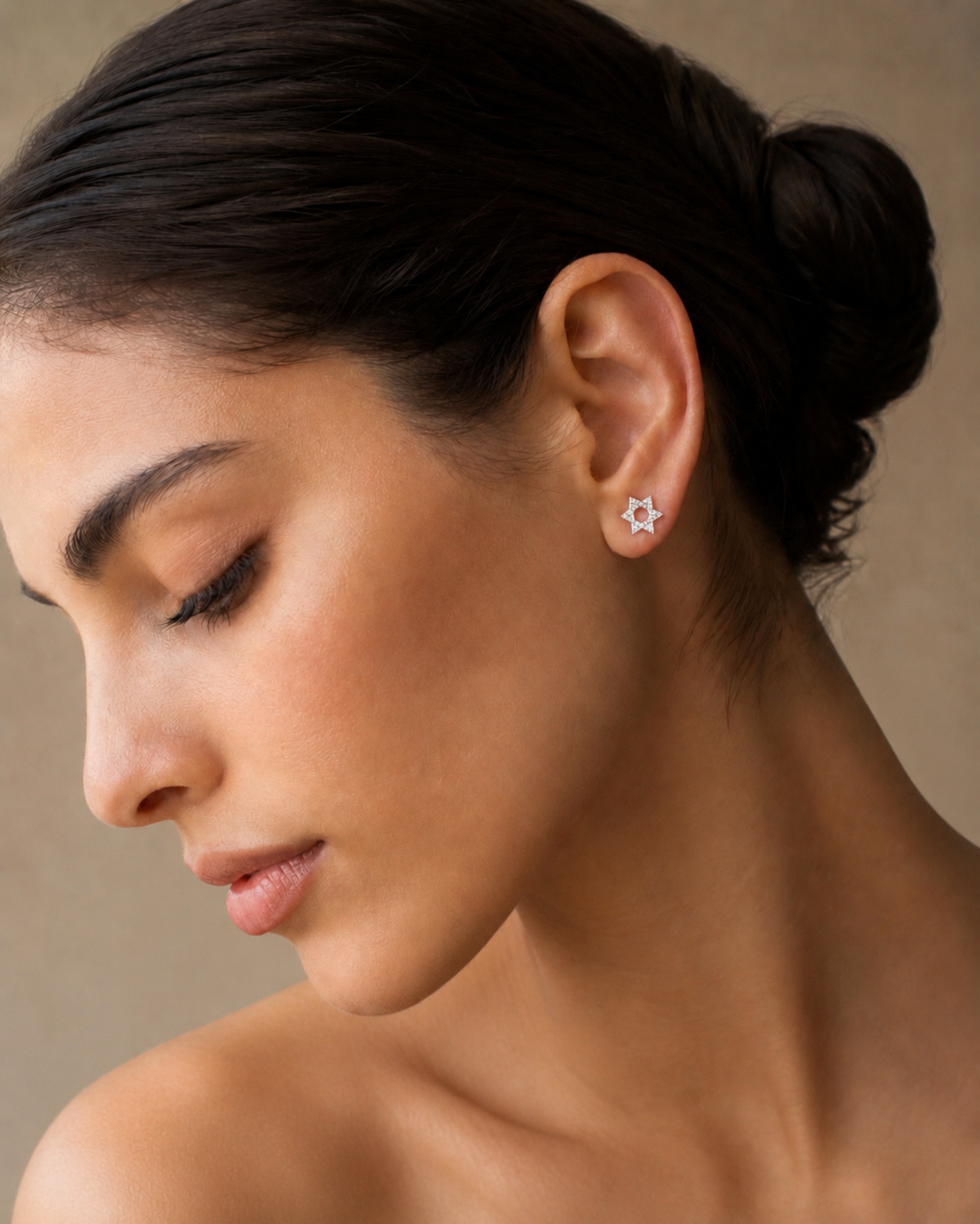 A close-up of a woman with dark hair in a bun wearing a small silver star-shaped stud earring, shown in profile against a soft beige background.
