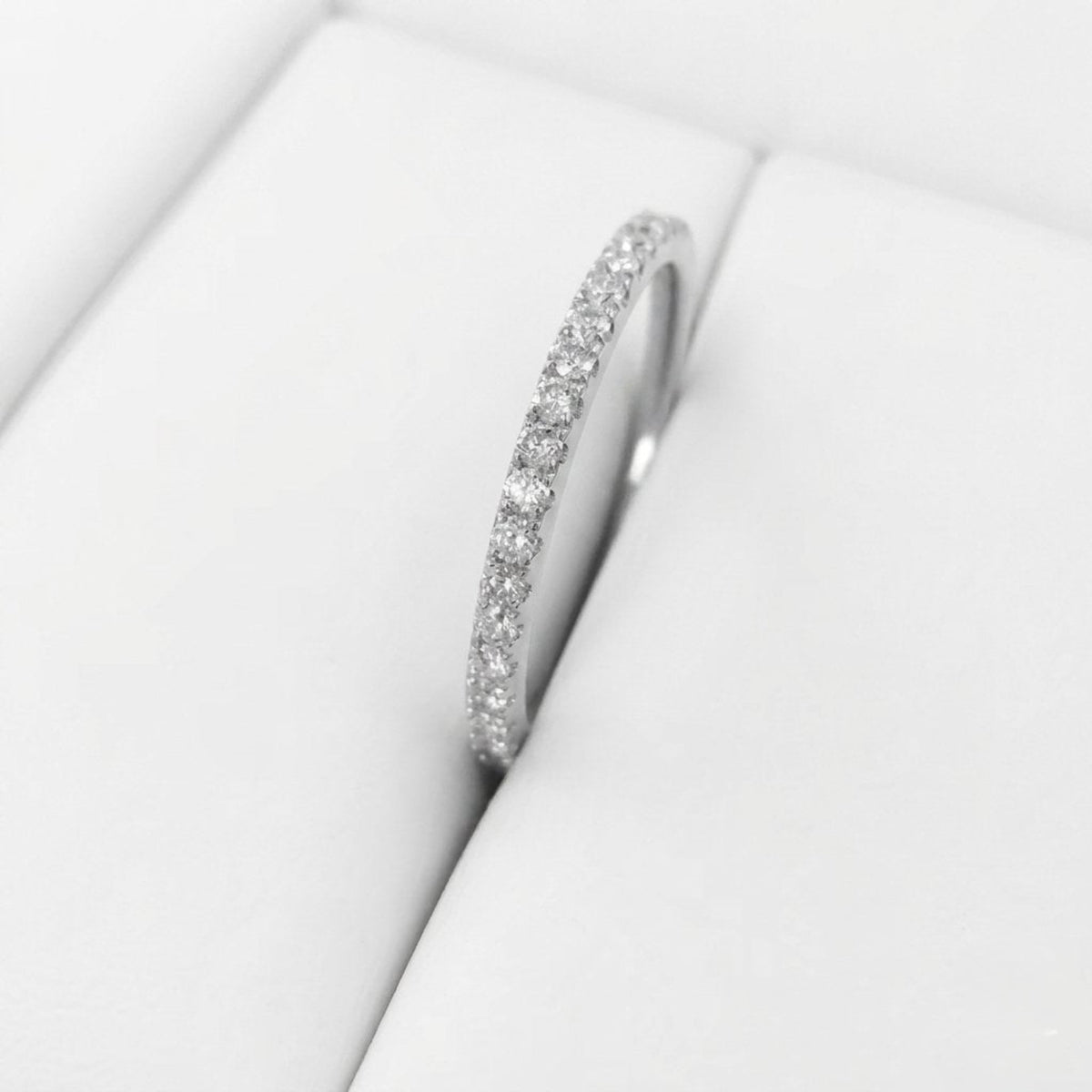 A silver-tone eternity ring set with small clear round diamonds, displayed on a white cushion inside a jewelry box.