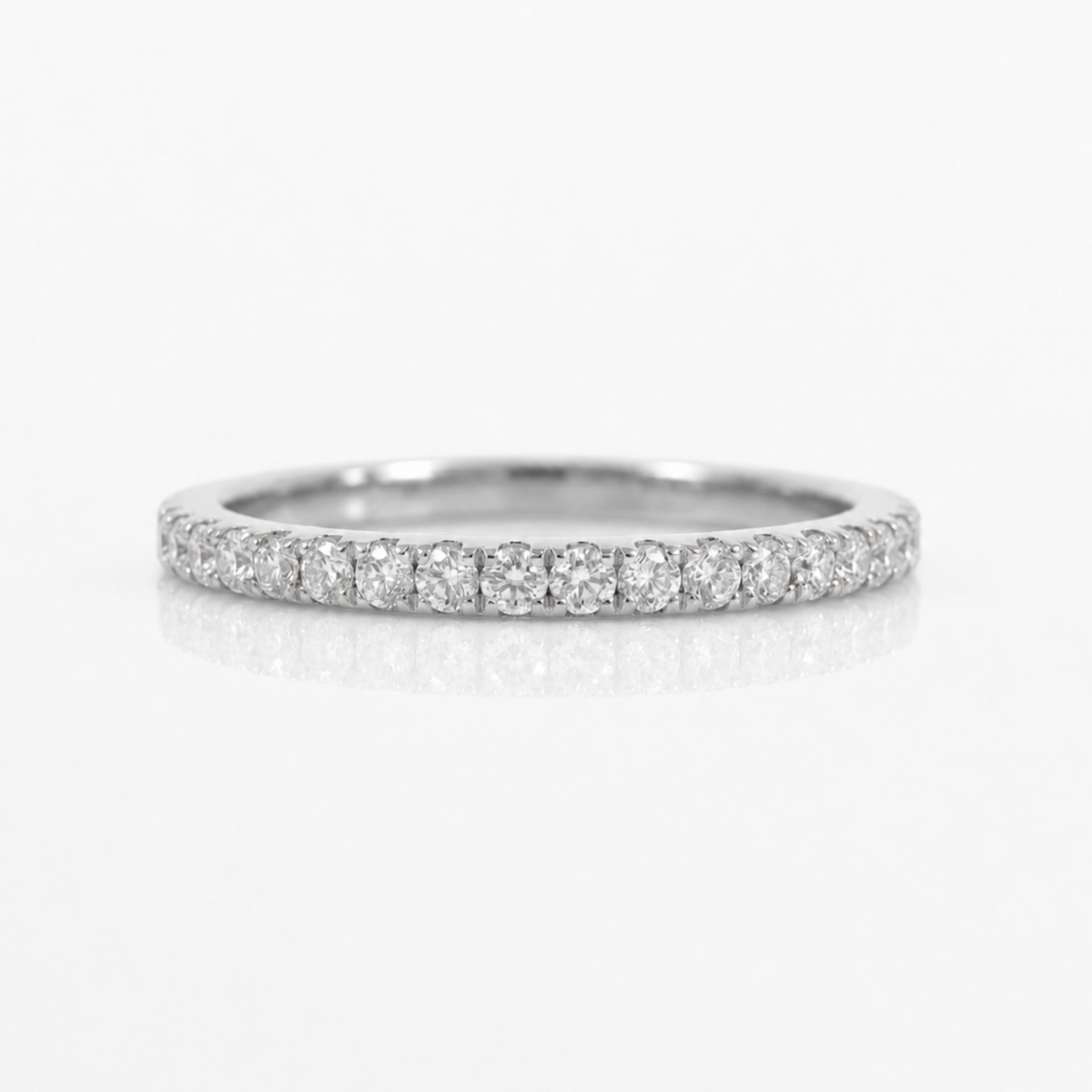 A thin diamond eternity band ring in a silver-toned metal, featuring a continuous row of round-cut diamonds set along the band, photographed on a clean white background with a soft reflection below.