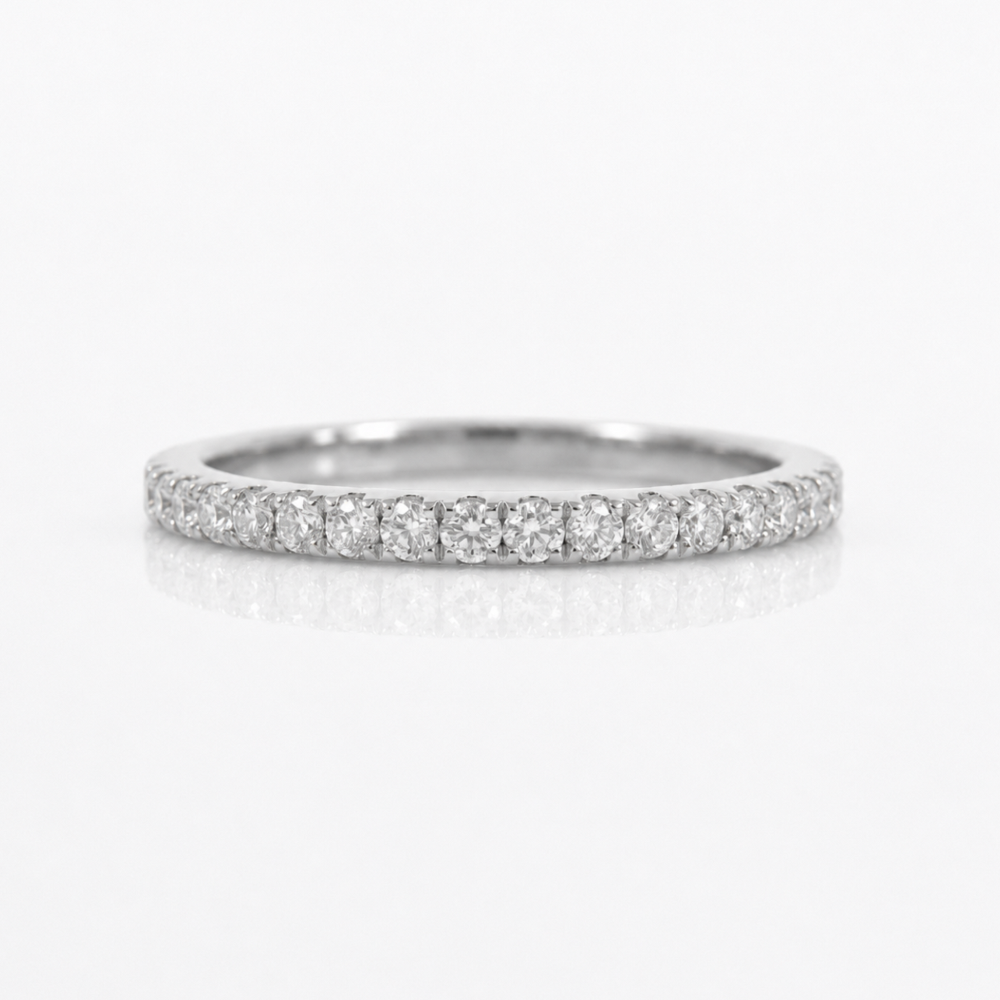 A thin diamond eternity band ring in a silver-toned metal, featuring a continuous row of round-cut diamonds set along the band, photographed on a clean white background with a soft reflection below.