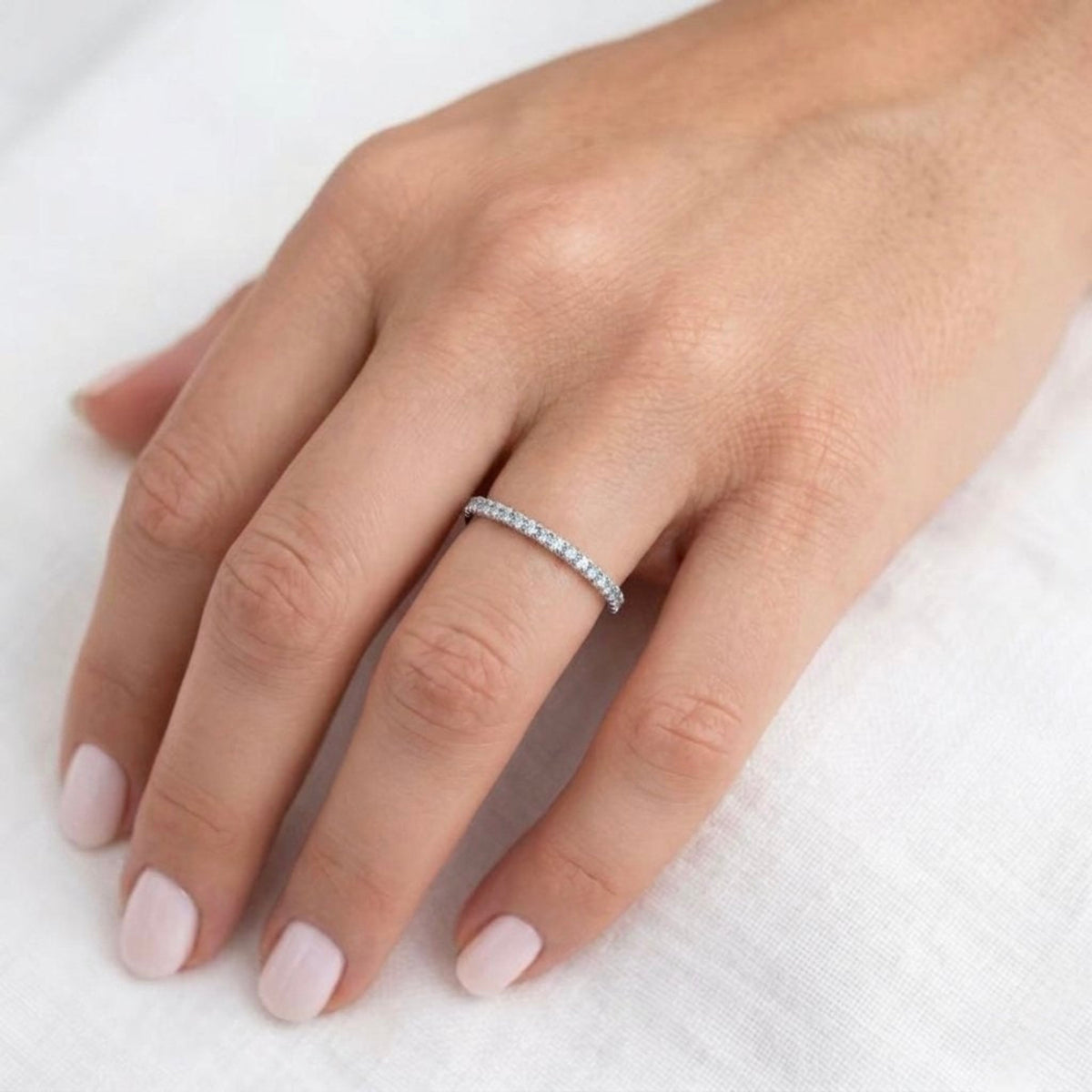 A delicate gold ring with a row of small clear diamonds worn on a person's ring finger, shown against a soft white textured fabric background.