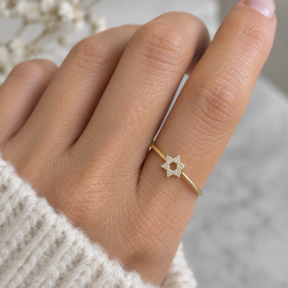 A close-up of a hand wearing a delicate gold ring with a small star-shaped diamonds cluster, set against a soft, blurred background.