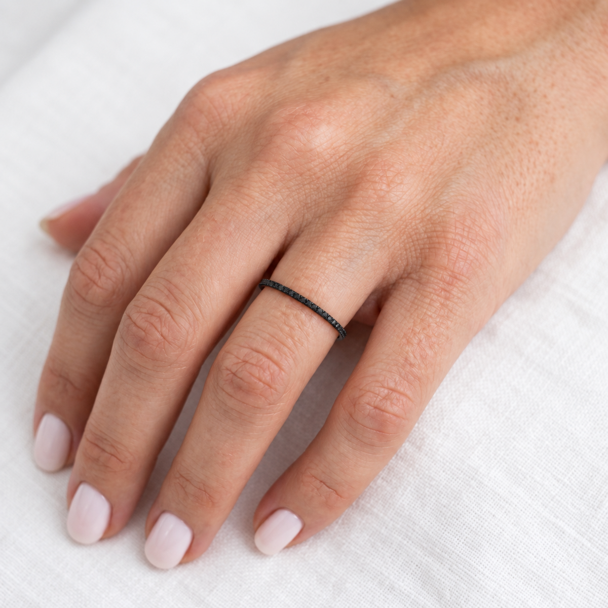 A close-up of a hand wearing a thin black black diamonds ring on one finger, resting on a white textured fabric background.