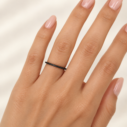 A close-up of a hand wearing a thin black ring with small black diamonds set along the band, against a soft light beige background.