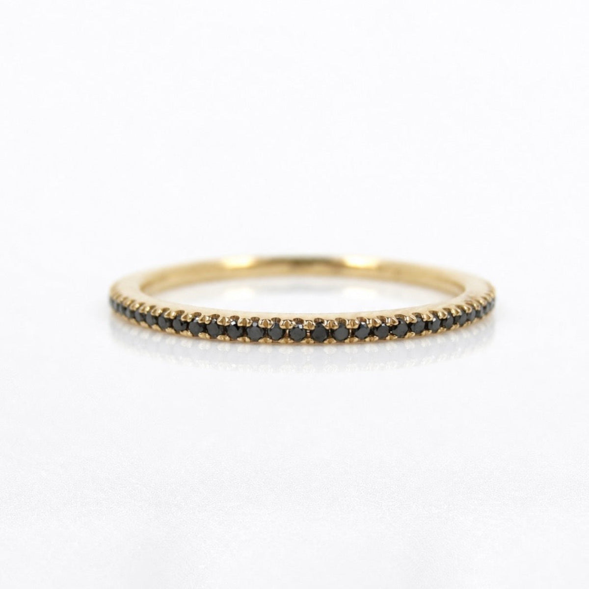 A delicate yellow gold eternity ring featuring a single row of small round black diamonds, photographed on a clean white background.