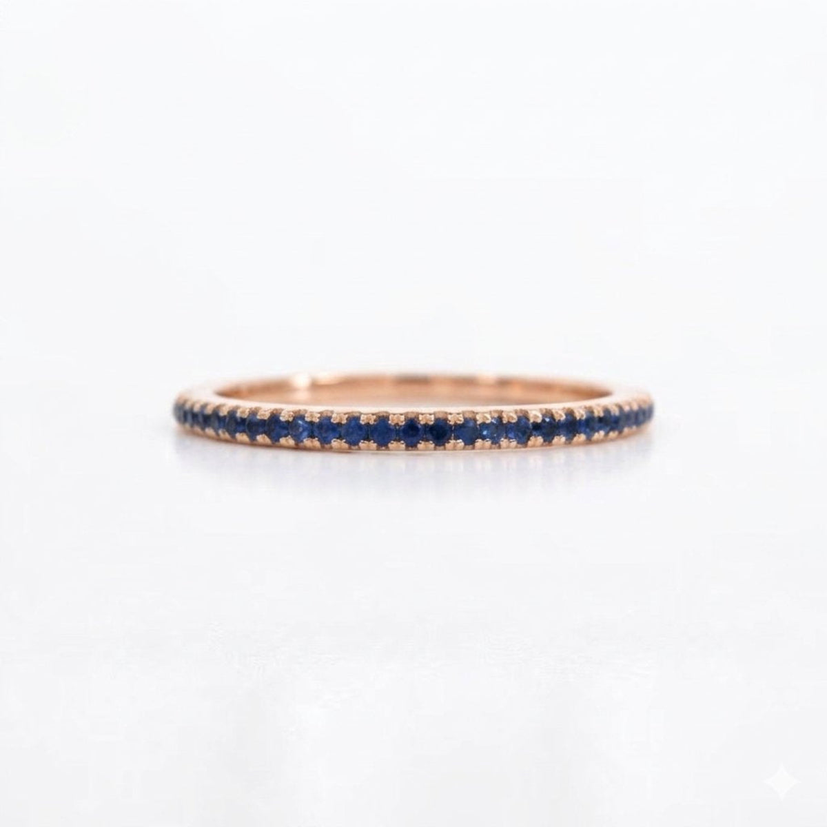 A delicate rose gold eternity ring featuring a row of small round-cut blue sapphires, shown on a clean white background.