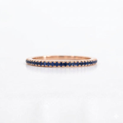 A delicate rose gold eternity ring featuring a row of small round-cut blue sapphires, shown on a clean white background.