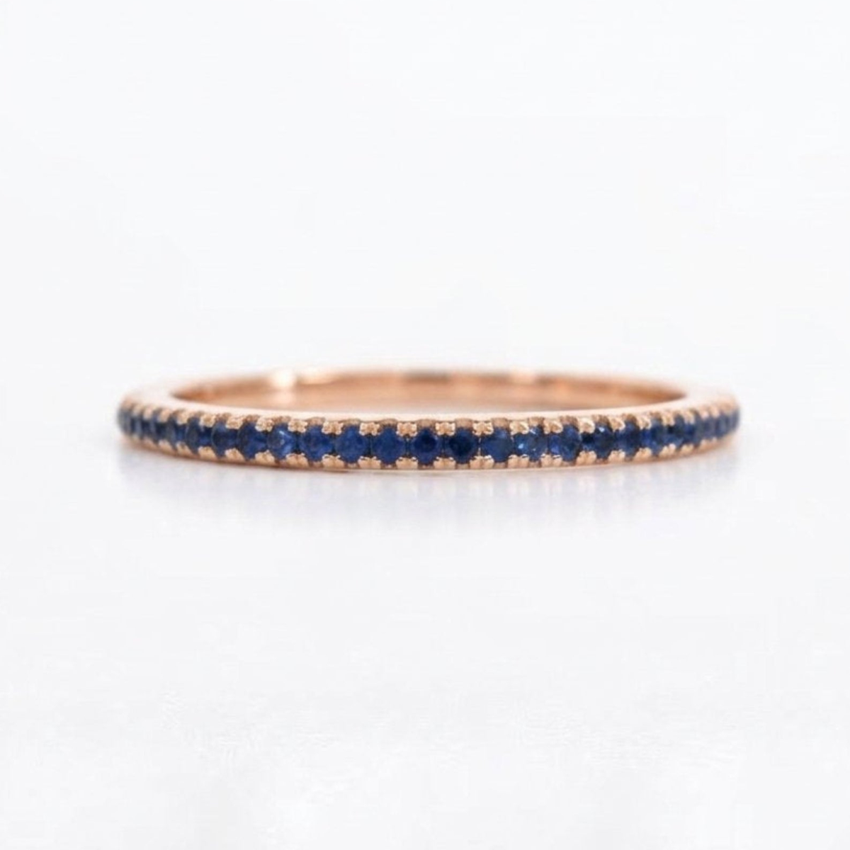 A rose gold eternity ring featuring a continuous row of round-cut deep blue sapphires, shown on a clean white background.