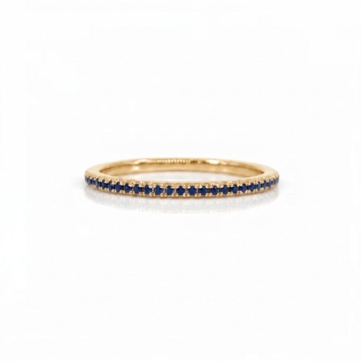 A delicate yellow gold eternity ring featuring a single row of round-cut deep blue sapphires, set against a clean white background.