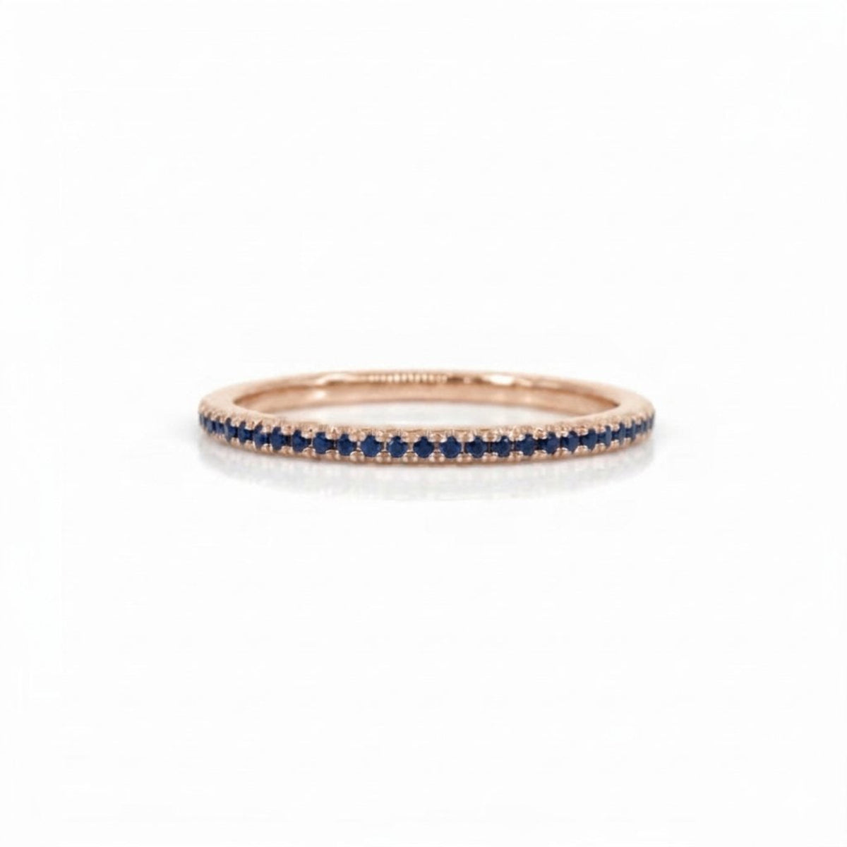 A delicate rose gold eternity ring featuring a single row of round-cut deep blue sapphires, photographed on a clean white background.