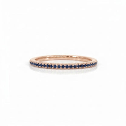 A delicate rose gold eternity ring featuring a single row of round-cut deep blue sapphires, photographed on a clean white background.