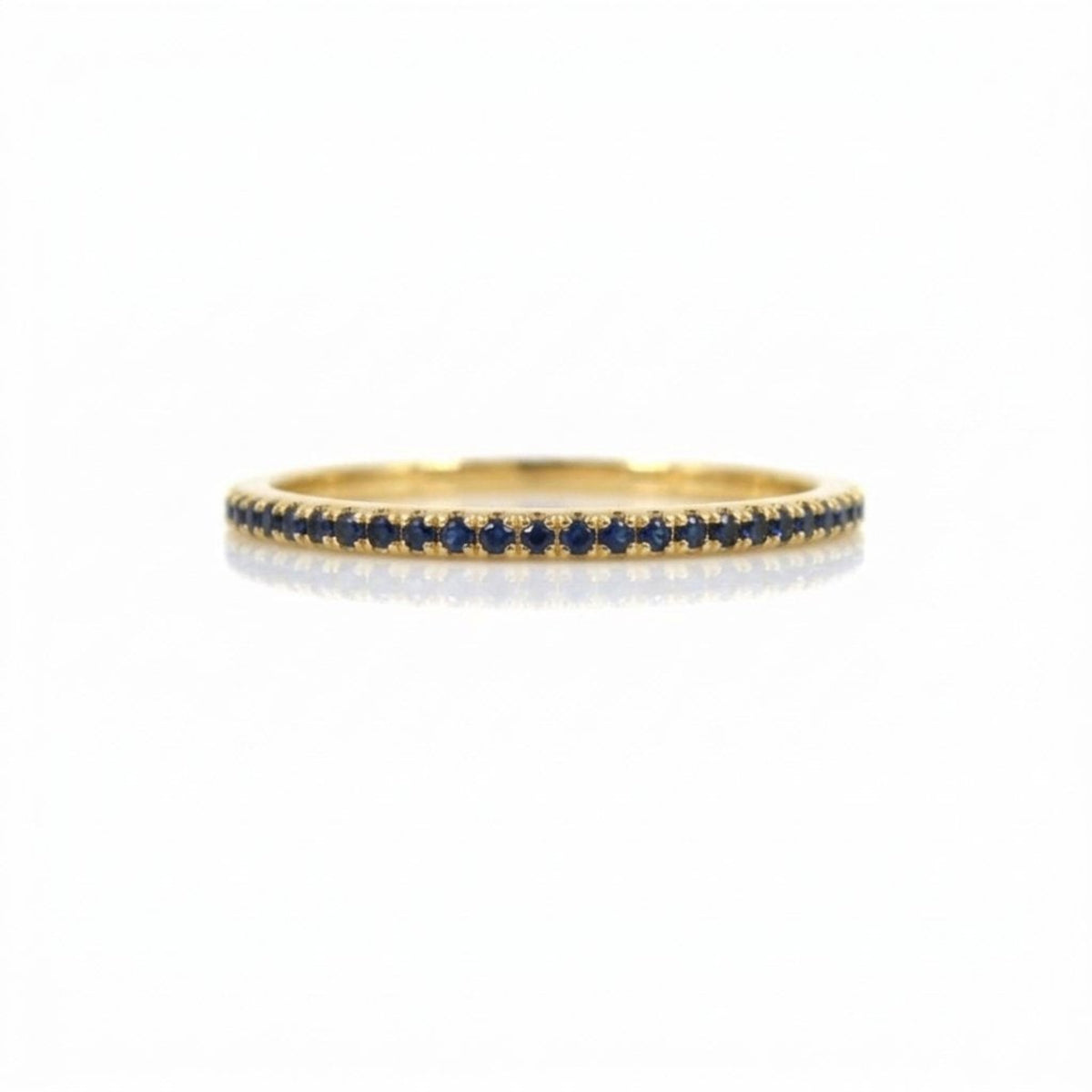 A thin yellow gold eternity ring set with a single row of small round dark blue sapphires, shown on a clean white background with a soft reflection.